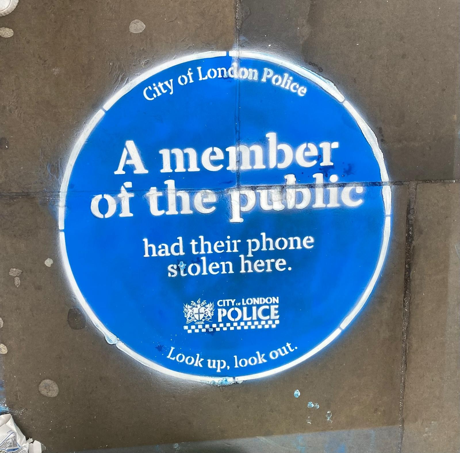 Blue circle spray painted on street. TEXT: A member of the public had their phone stolen here. Look up, look out.